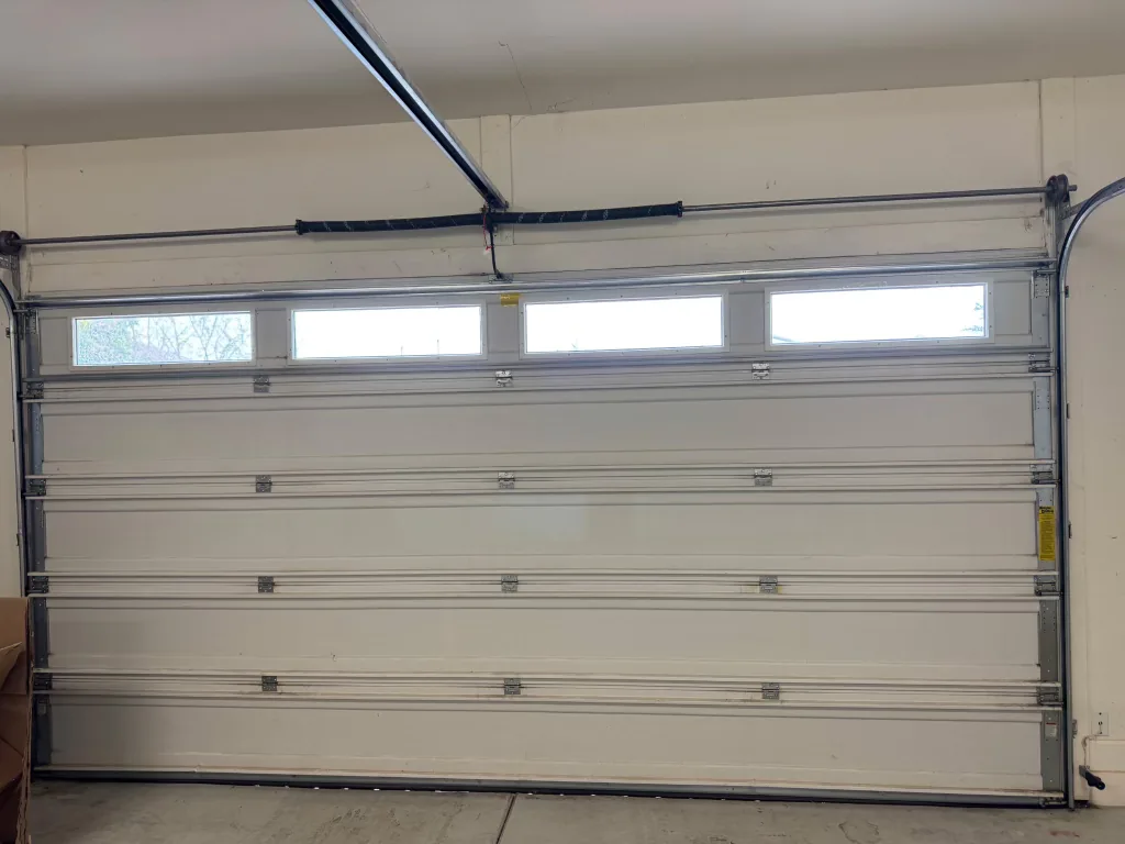 Installed white sectional garage door seen from inside the garage, with a row of window inserts in the top panel and the track, hinges and torsion shaft in view