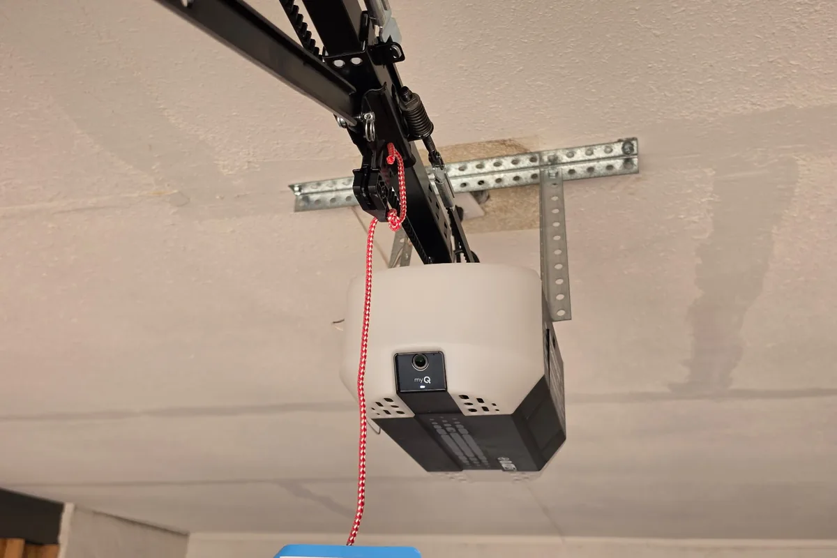 Ceiling-mounted garage door opener with its rail, mounting strut and red manual release cord — the hardware checked over during an annual service